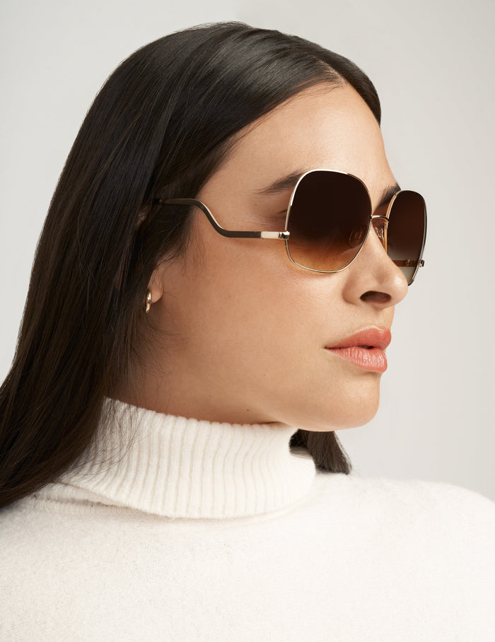 Oversized square gold-tone metal sunglasses with gradient brown lenses and drop temple detail, worn by a model in profile.