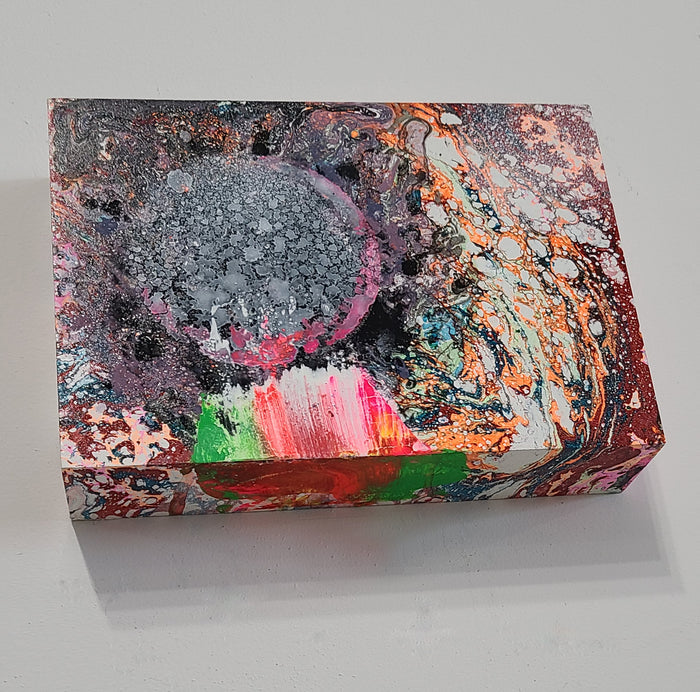 The artwork 'Phantom Planet' features a cosmic-inspired abstract composition with a textured gray spherical form surrounded by swirling patterns in pink, orange, and turquoise. Bold strokes of neon green and red create dynamic movement across the wooden surface, executed in gouache, spray paint, and conte crayon by Millie Benson.